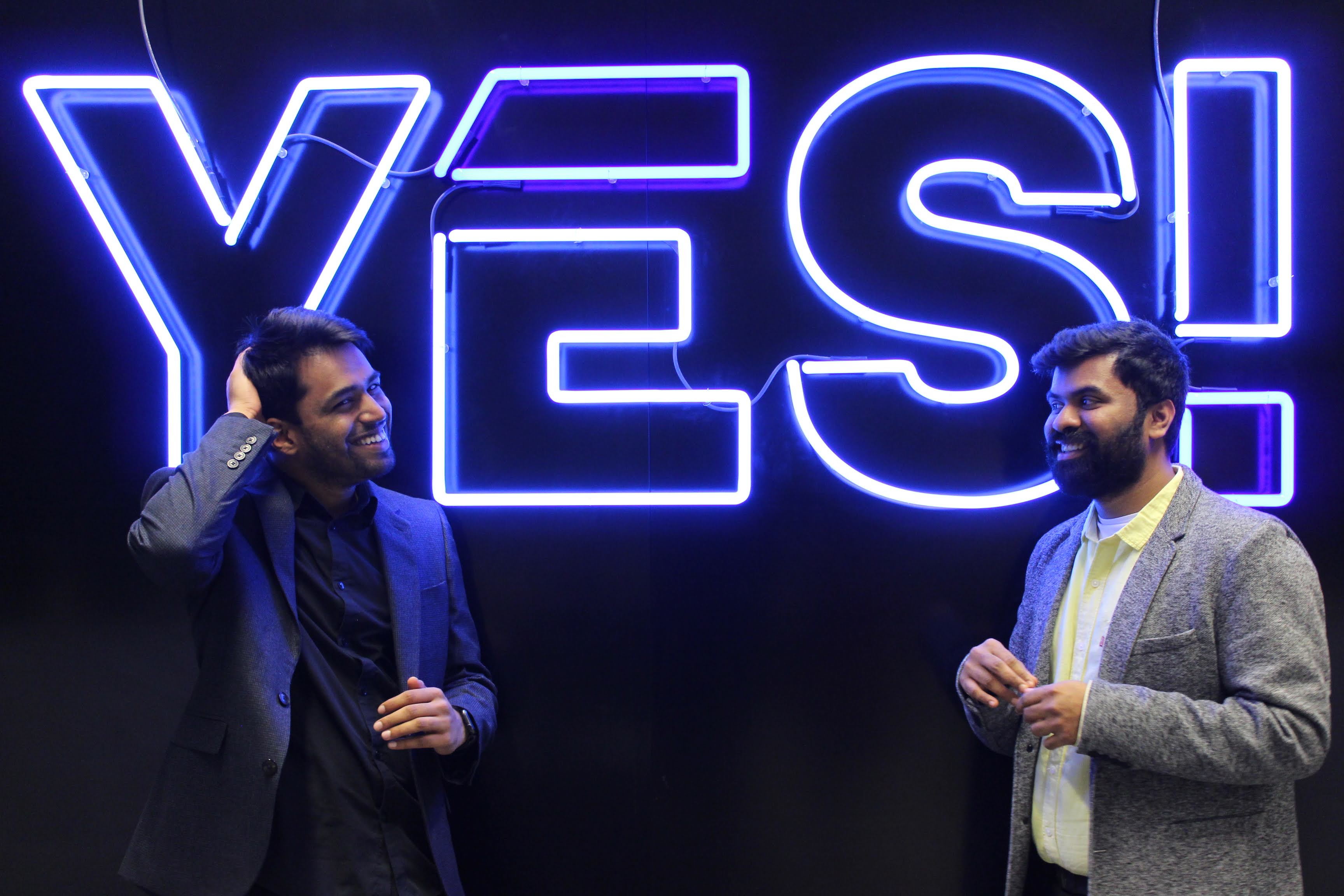 Founders Karthik Mahadevan and Karthik Kannan smile while posing in front of a big blue neon sign which reads YES!, the logo of Envision's hub.