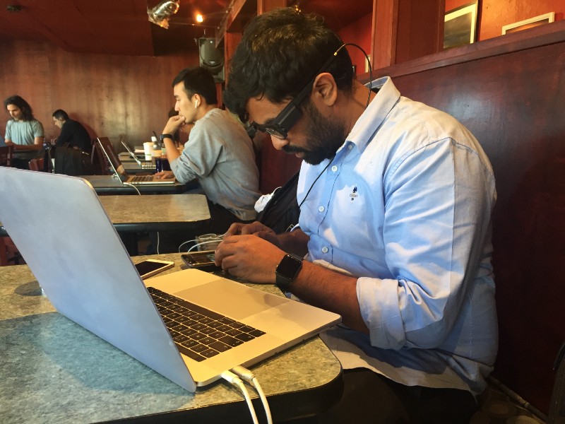 Co-founder Karthik Kannan is sitting at a cafe, wearing the prototype glasses while he works on his laptop and smartphone.