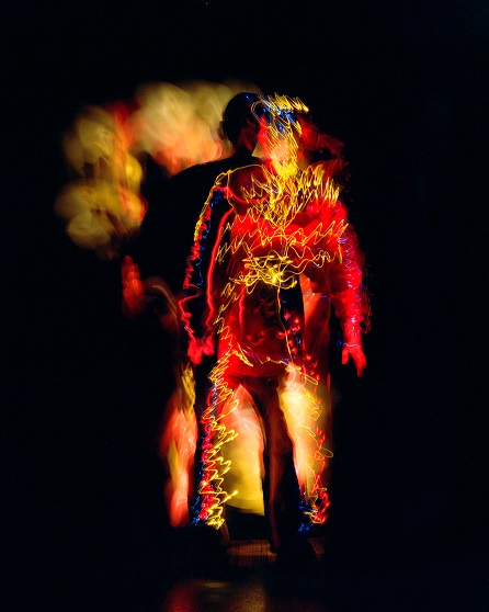 The figure of a man is seen on a black background. The man is covered in wiggly yellow, red and some blue colours.