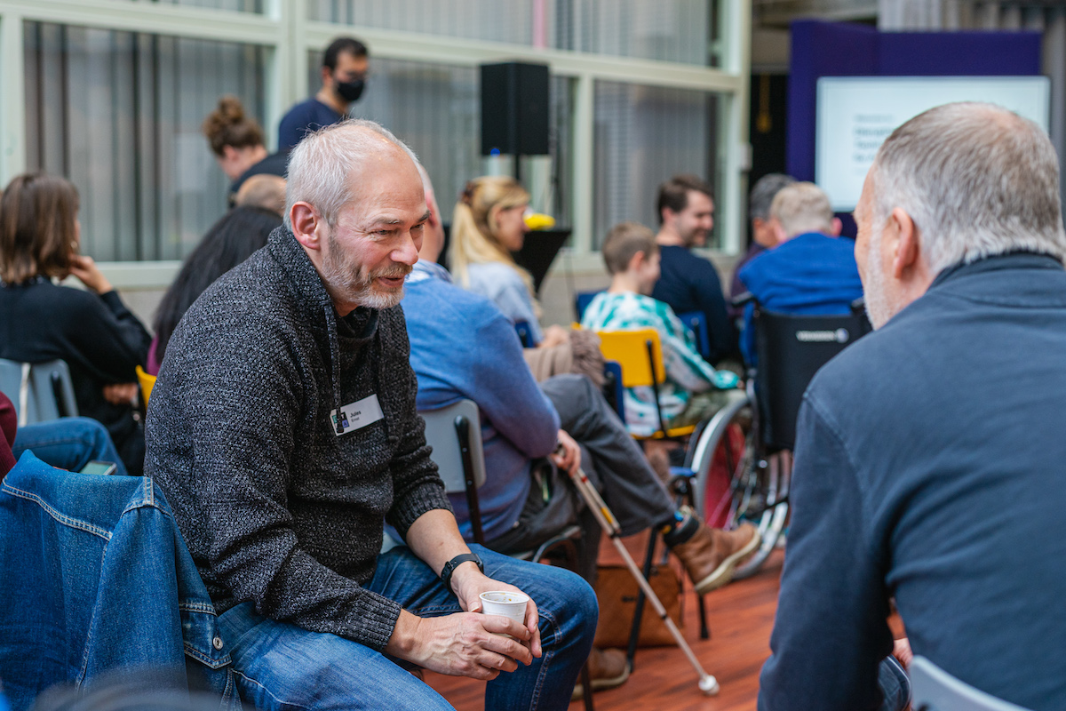 Two people talking to each other. In the background there is a group of people sitting and waiting to watch a presentation.