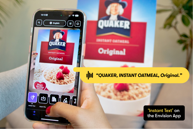 A person is scanning a Quaker Instant Oatmeal box by holding their phone over it, using the Envision App to read it. It shows a yellow speech bubble which means the content of the document is spoken out. Bottom-right text box states: Instant Text on the Envision App.
