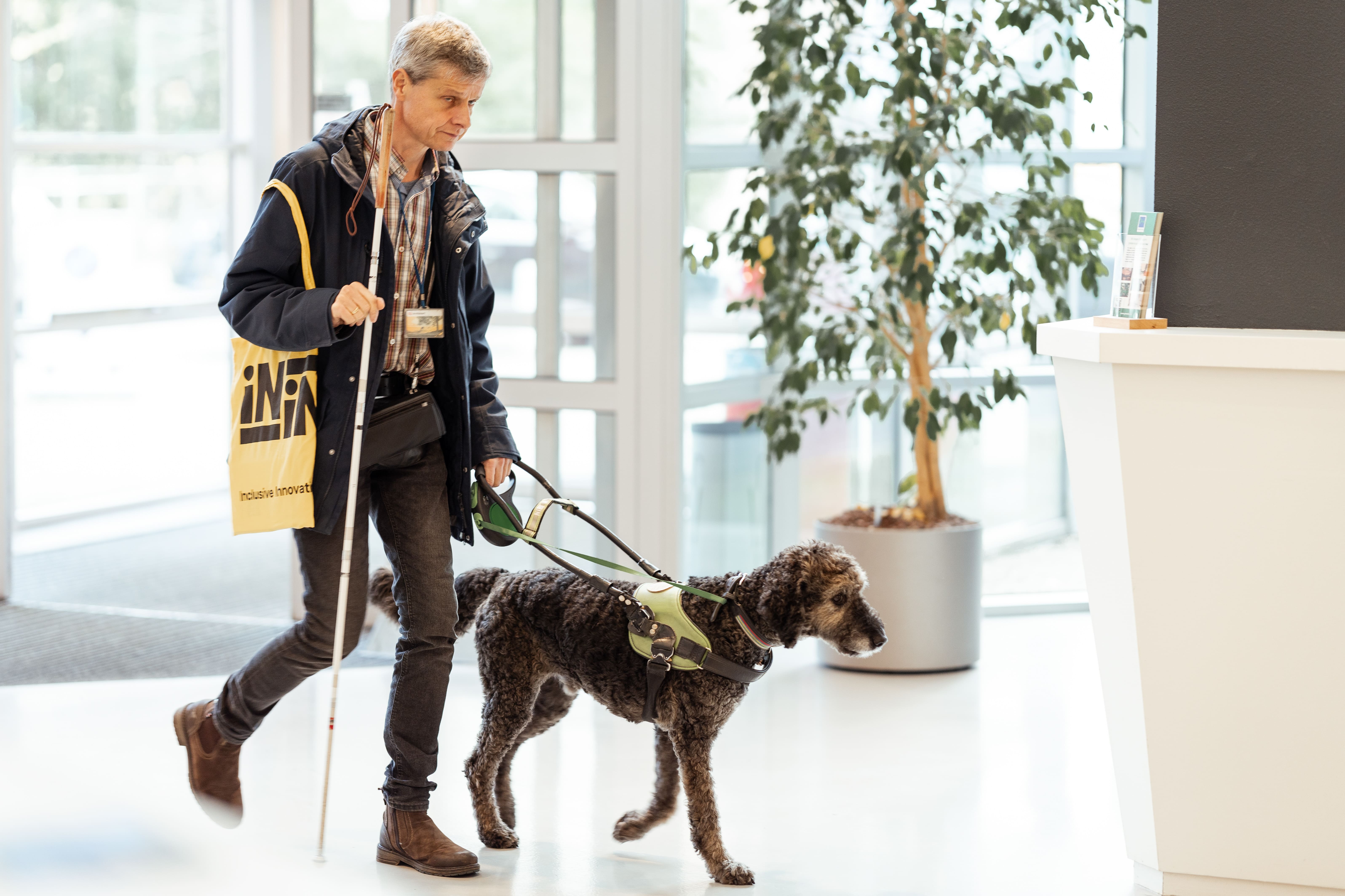 Man walking indoors with a white cane and a guide dog wearing a harness.