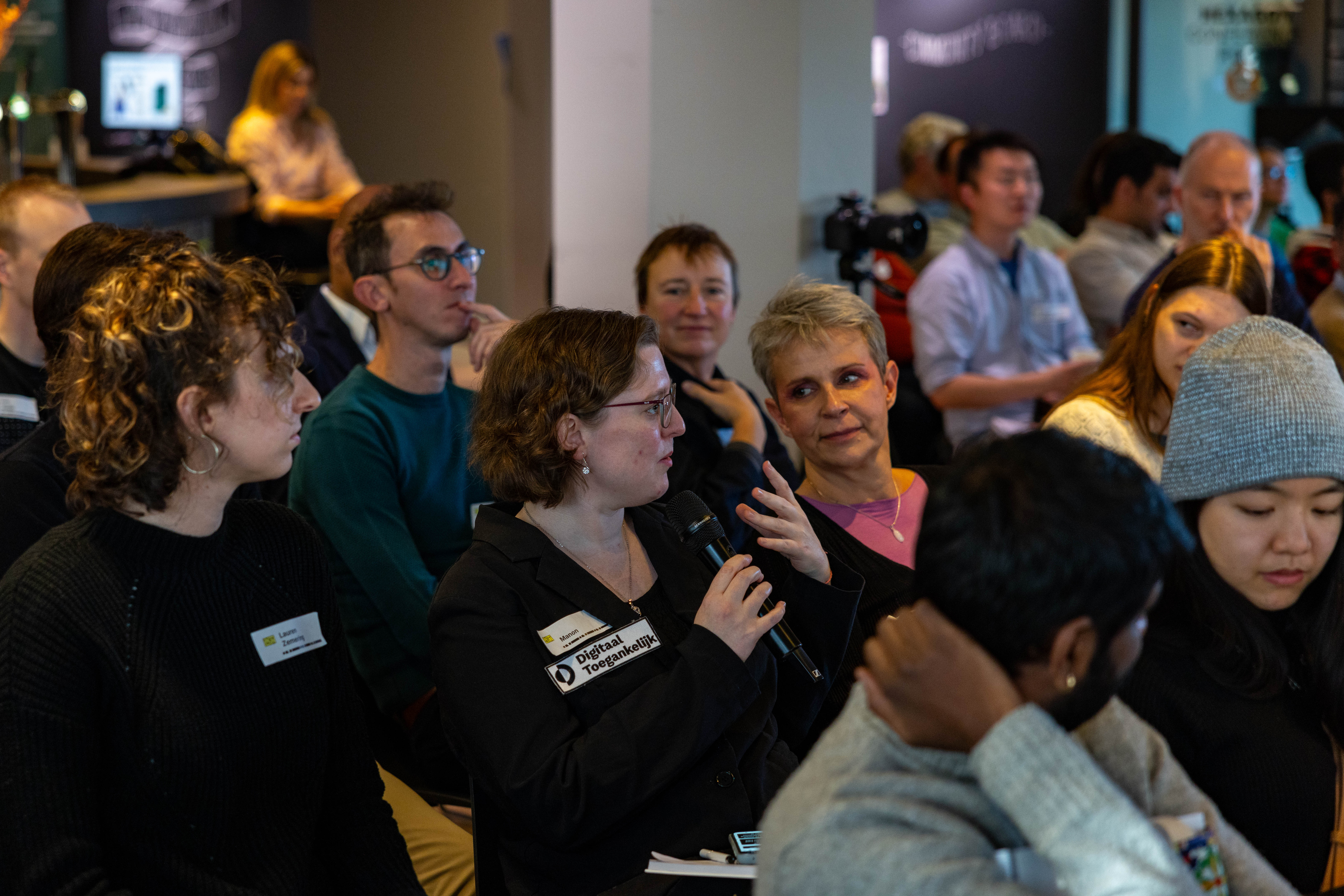 A group of people seated in an indoor setting, engaging in a discussion or event. A woman in the center, wearing glasses and holding a microphone, is speaking while others listen attentively. Name tags are visible on some attendees. The background shows more participants, some blurred, in a casual environment with subdued lighting. 