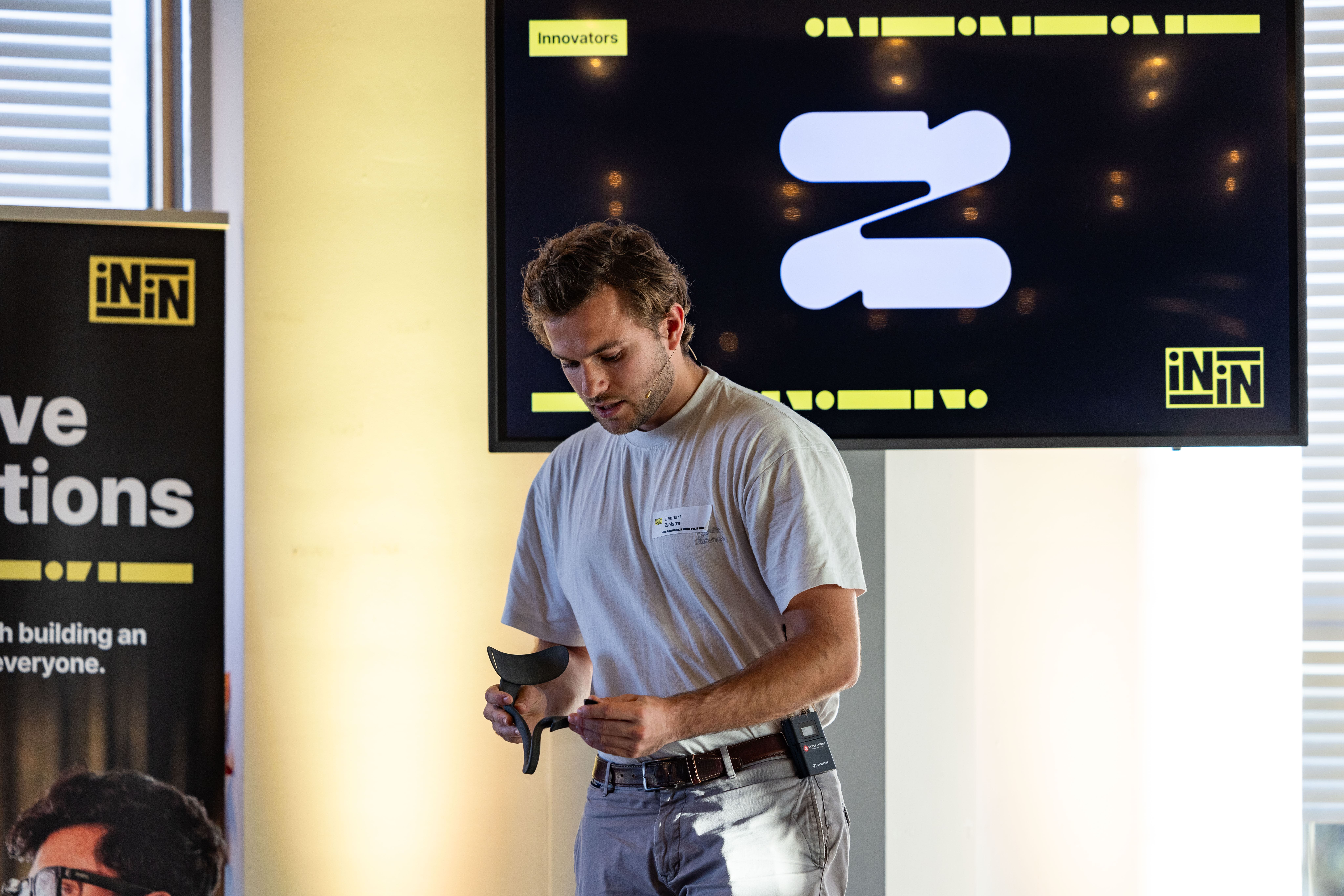 A man wearing a white t-shirt and a headset microphone stands in a presentation setting, holding a black ergonomic object in his hands. Behind him, a large screen displays a minimalist logo with abstract shapes and the text "Innovators" in yellow at the top. To the left, part of a banner with the words "Innovative Solutions" is visible.
