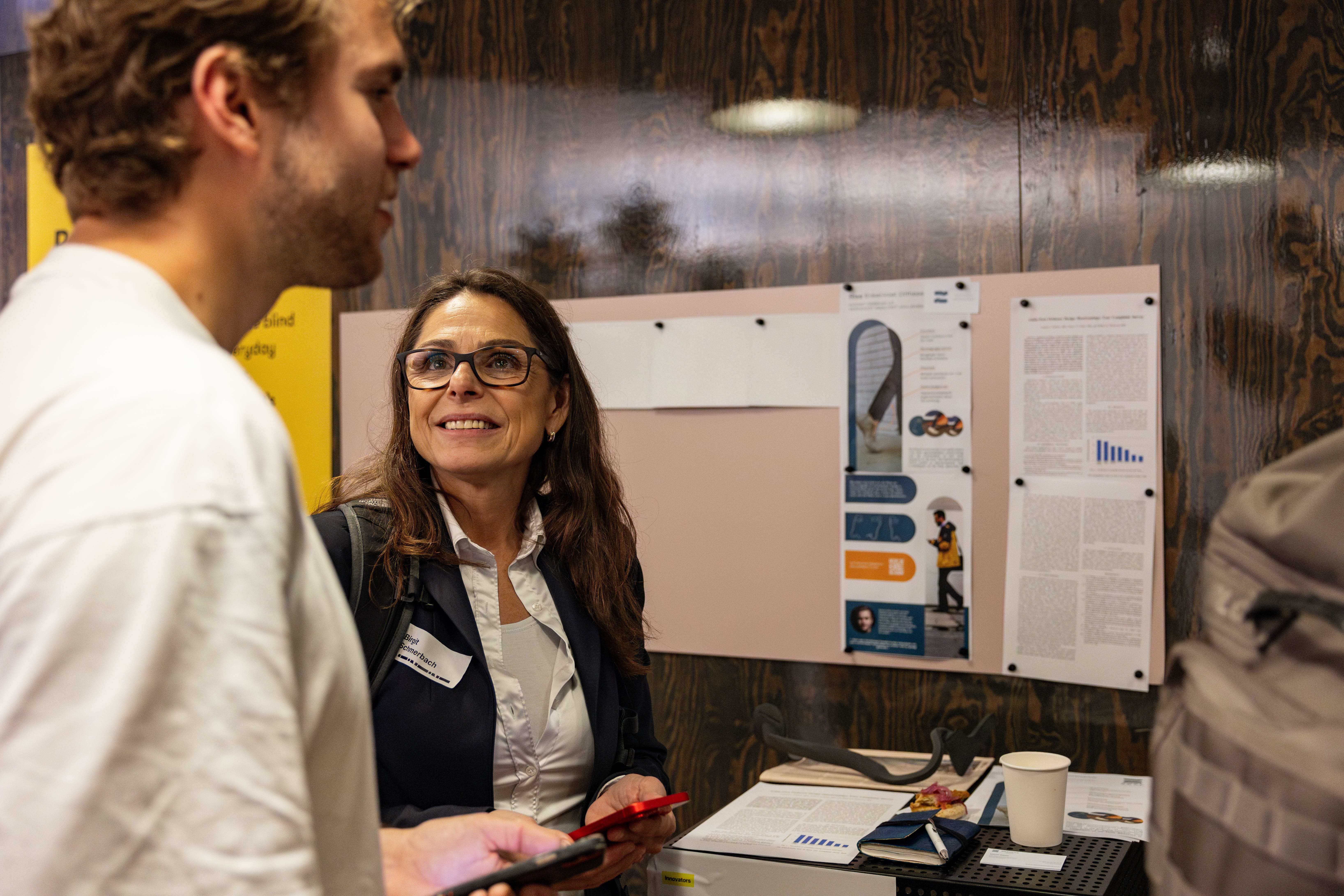 A woman wearing glasses, a white shirt, and a dark blazer smiles while conversing with a man in a white shirt, who is partially visible in the foreground. The interaction takes place in front of a bulletin board displaying various printed materials, including charts, graphics, and articles. A table in front of the board holds papers, a white paper cup, and other small items. The wall behind the board is dark wood-paneled.
