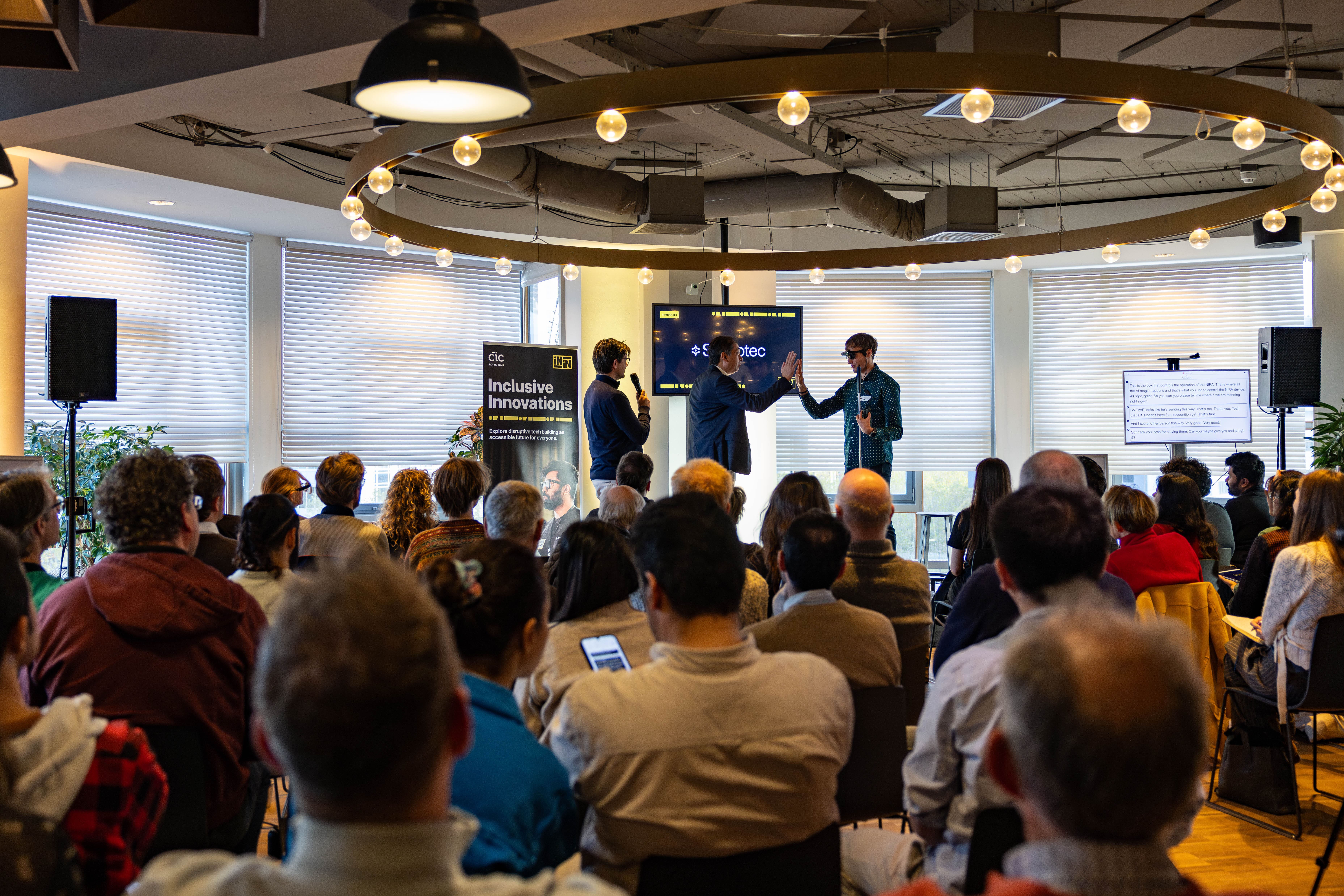 An audience seated in a brightly lit room watches a presentation. Three speakers stand at the front under circular string lights. Two of the speakers, one in a navy jacket and the other in a green patterned shirt and sunglasses, exchange a high-five while holding microphones. A screen behind them displays a presentation, and a banner nearby reads "Inclusive Innovations." Additional text is visible on a screen to the right, and potted plants decorate the space.
