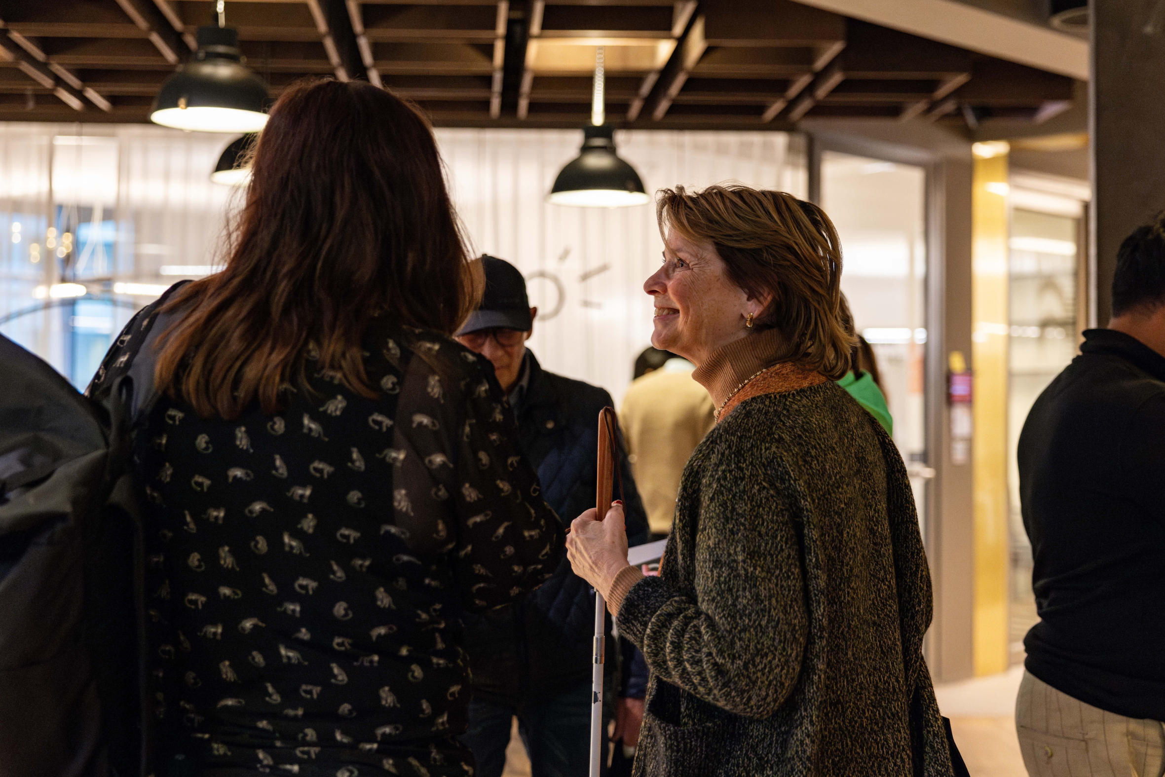 A group of people gathered in a well-lit indoor space. In the foreground, a woman wearing a patterned black top with animal prints is seen from behind, facing another woman who is smiling and holding a white cane. The smiling woman wears a brown turtleneck sweater and a green cardigan. Other individuals are visible in the background, standing and conversing near modern light fixtures and glass walls.
