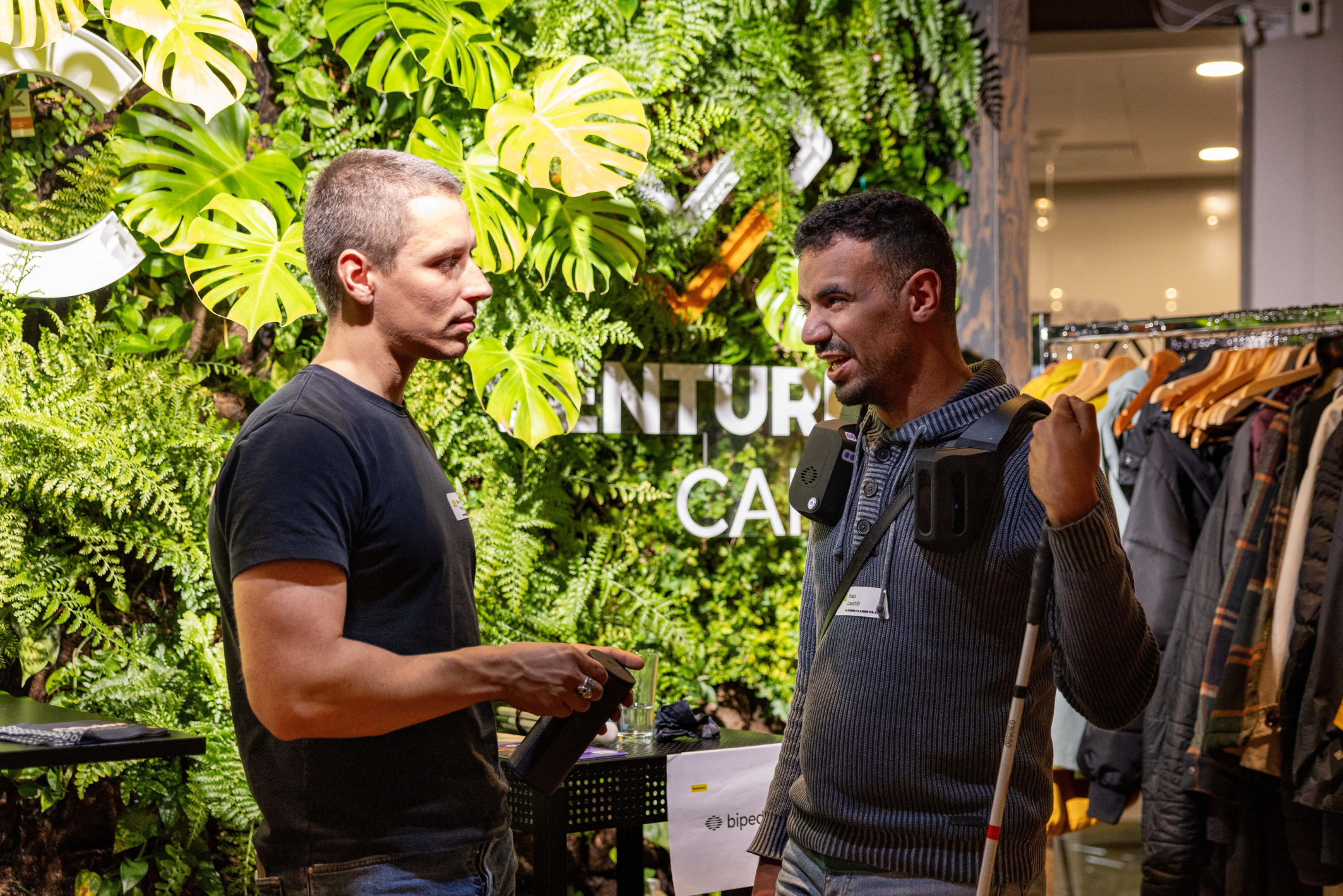 Two men having a conversation in front of a vibrant green wall decorated with large monstera leaves and ferns. The man on the left, wearing a black t-shirt, holds a black cylindrical object and looks attentively at the other man. The man on the right, dressed in a gray sweater with a tactile device hanging from his shoulders, is smiling and holding a white cane. In the background, there is text on the wall partially visible, clothing racks, and soft lighting.
