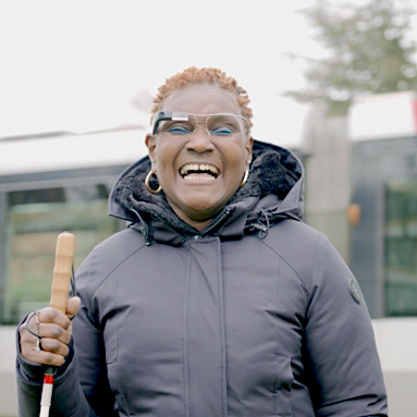 Front view of a person holding a white cane and wearing Envision Glasses outside.