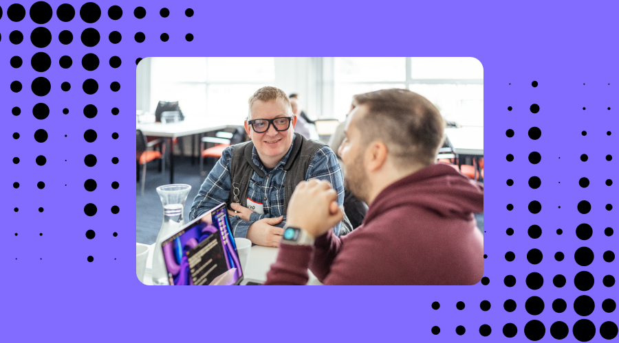 Two men engaging in a conversation at an office setting. The man facing the camera is smiling and wearing Envision Smart Glasses, along with a plaid shirt and a backpack. The other man, seen from the back, is wearing a burgundy shirt and gesturing with his hand. The surrounding area appears to be a bright and modern workspace, and the image is framed with a border featuring a pattern of black dots on a purple background.