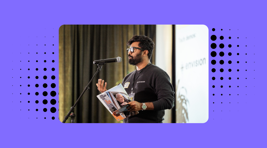 Karthik Kannan, the CTO and co-founder of Envision, is giving a demo. He is standing at a microphone and gesturing with one hand, holding a magazine or brochure with the other. The background includes a blurred projection screen with text related to the event.